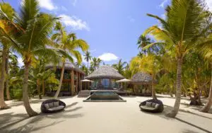 Outdoor lounge area of a French Polynesian villa, featuring a small pool and two woven sofas among palm trees on a private beach.