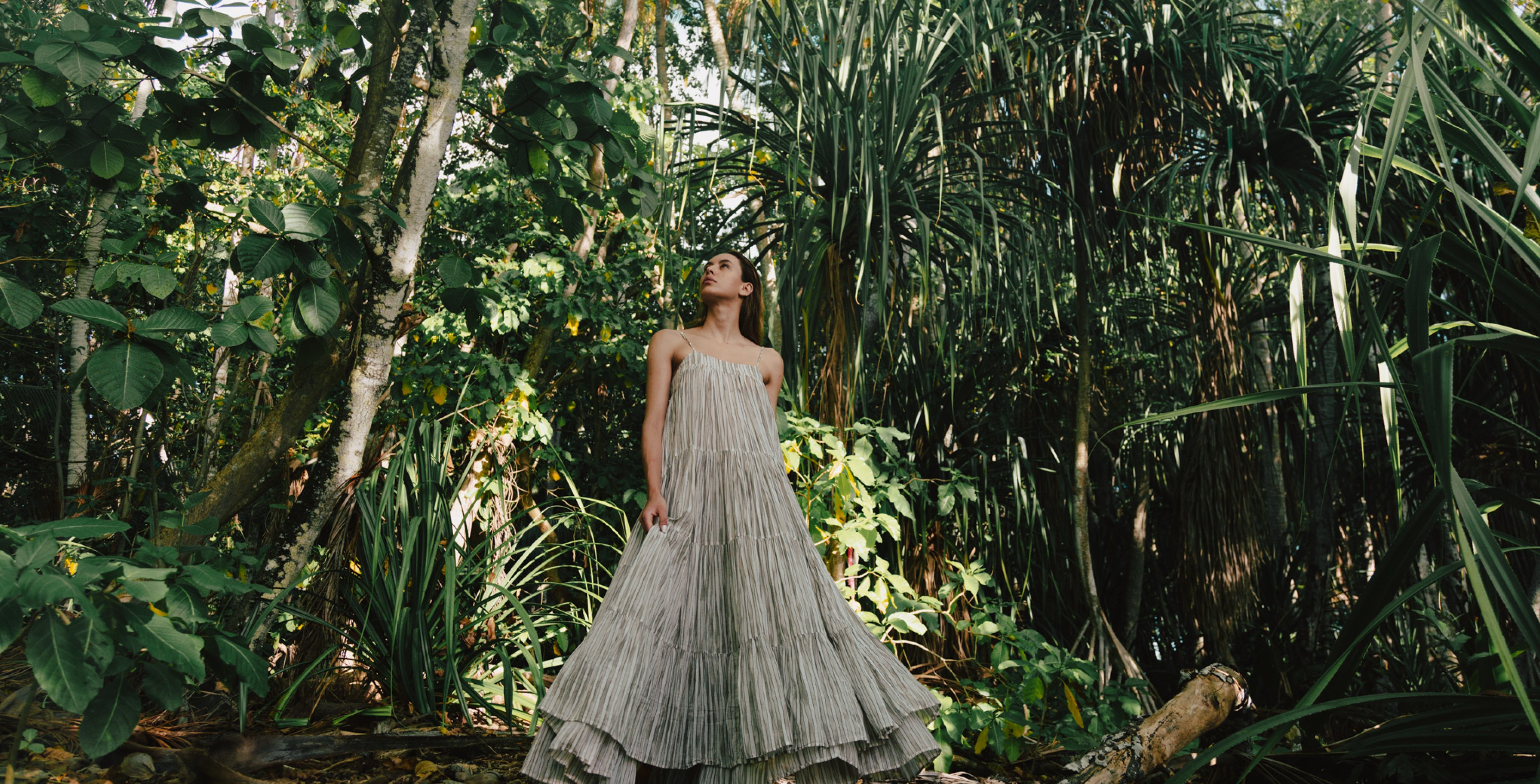 Woman in an off-white maxi dress, looking upwards while surrounded by lush greenery at our resort in Tahiti