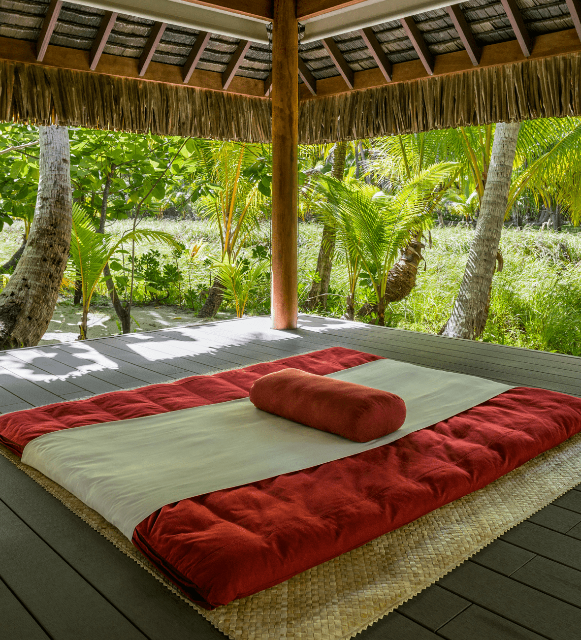 Red and white mattress on the ground of a hut surrounded by palm trees